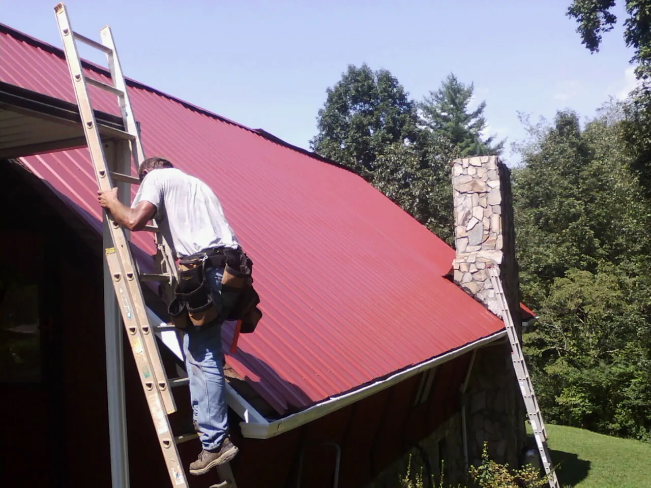 Roofer on a ladder beside a red metal roof and stone chimney — Soffit & Fascia Repair by Roofing Brothers in Mountville